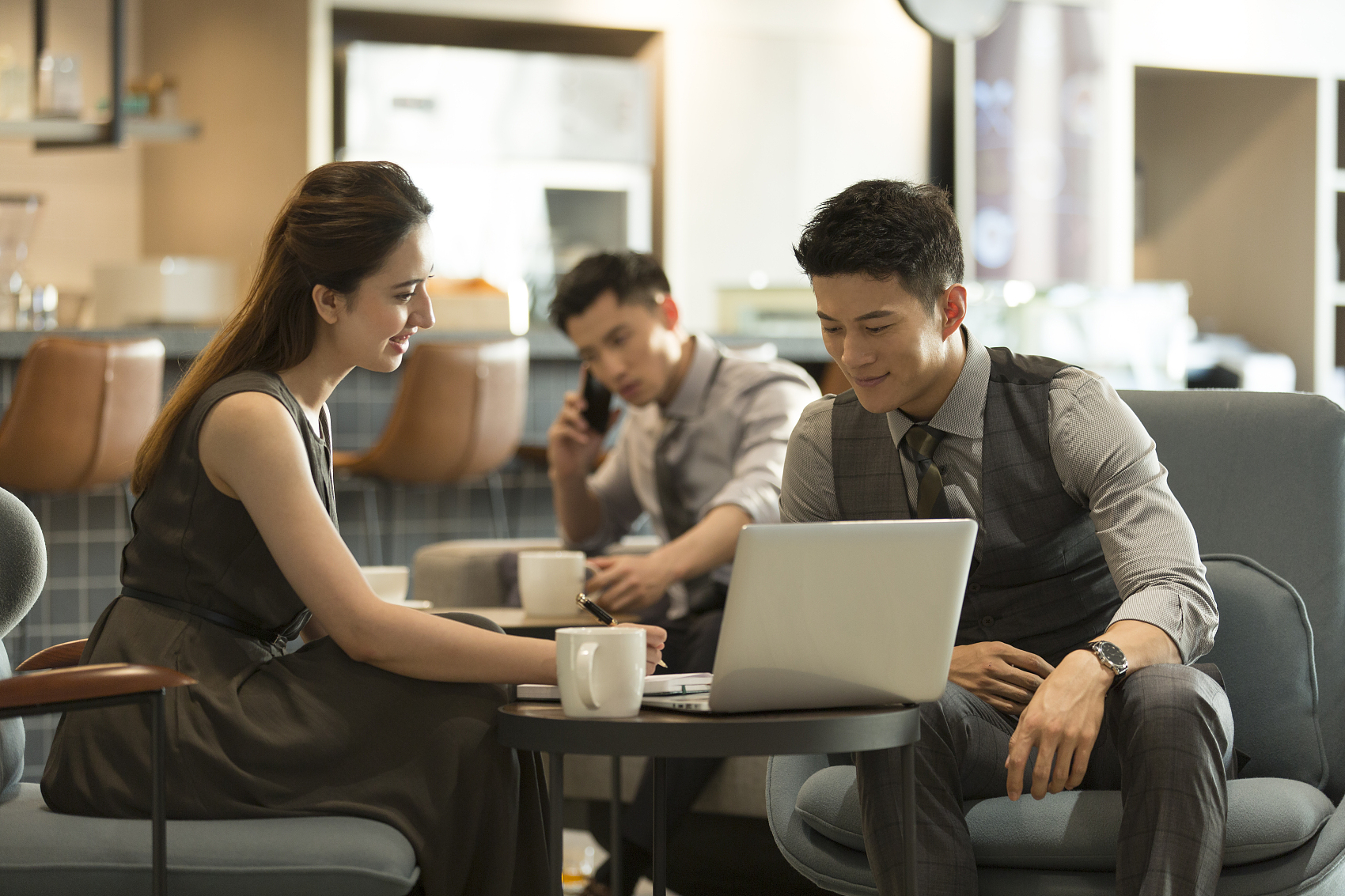 青年商务男子和青年女助理在办公室休闲区喝咖啡使用笔记本电脑探讨沟通工作